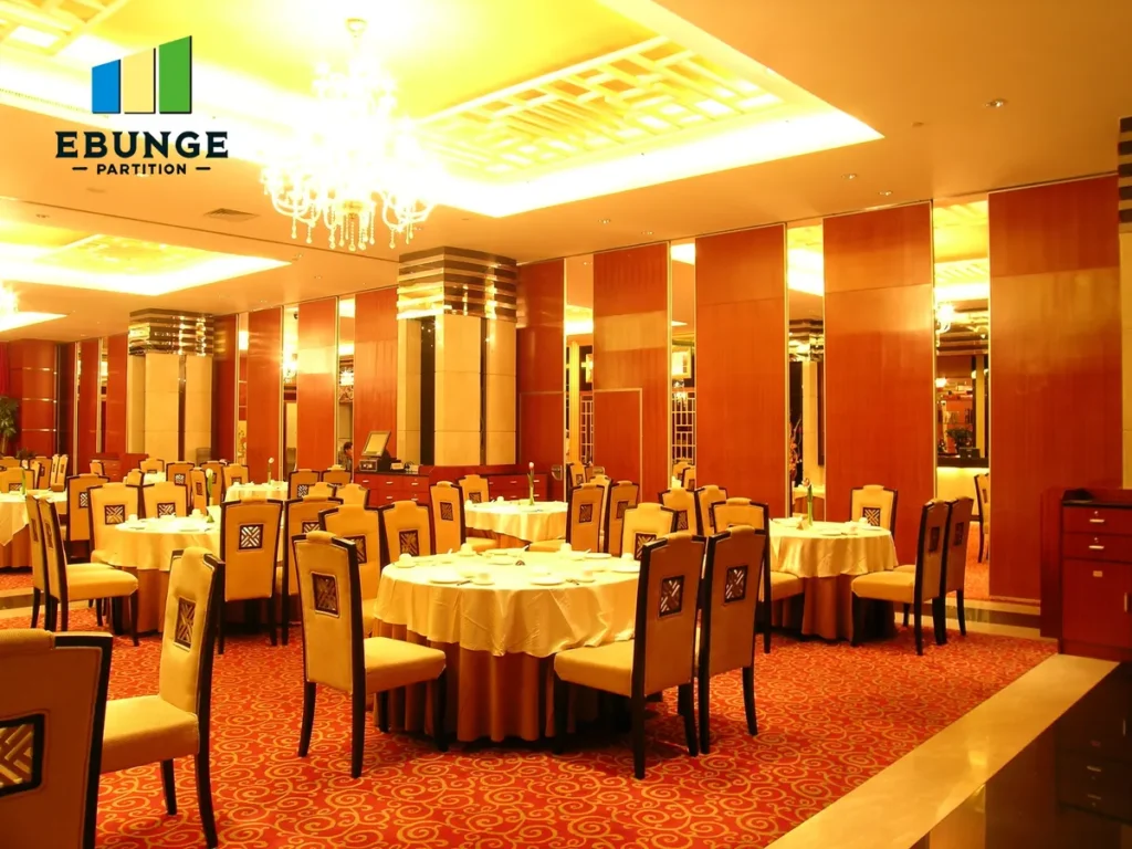 Banquet hall set for an event with round tables, beige chairs, white tablecloths, warm yellow lighting, chandeliers, and EBUNGE PARTITION logo top-left.