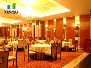 Banquet hall set for an event with round tables, beige chairs, white tablecloths, warm yellow lighting, chandeliers, and EBUNGE PARTITION logo top-left.